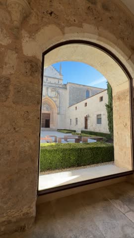 View through a broad stone arch toward the Gothic church facade, landscaped courtyard and benches at Miraflores Charterhouse in Burgos, Spain.