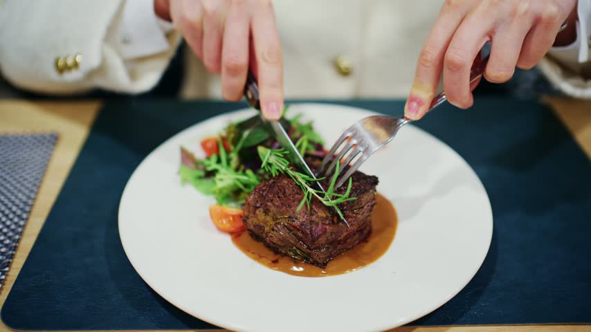 Person using knife and fork to slice juicy beef tenderloin steak served with fresh green salad and sauce at elegant dining table.