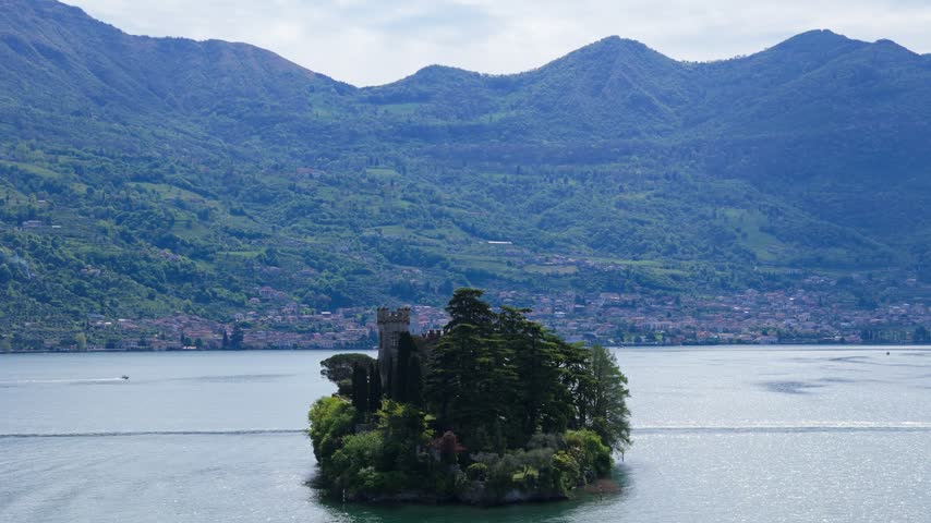 Aerial drone zoom in toward Isola di Loreto, a small private island with a historic castle surrounded by trees in the blue waters of Lake Iseo, Lombardy, Italy, with mountains in the background.