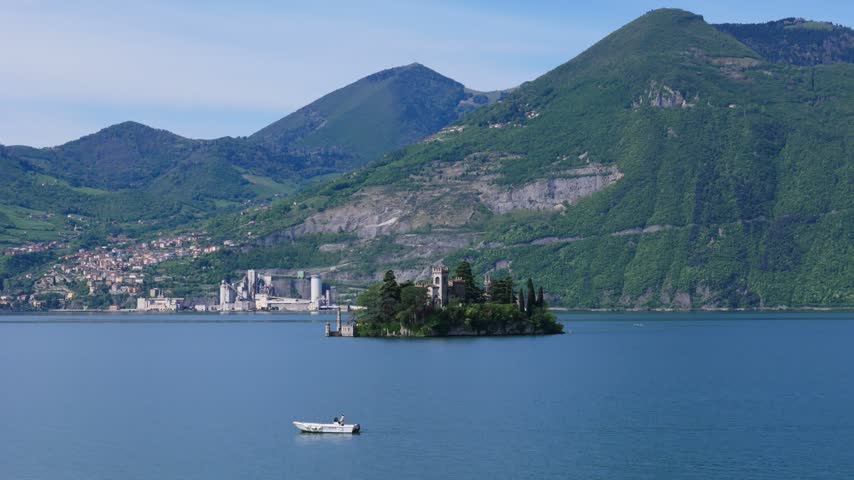 Aerial drone zoom in toward Isola di Loreto, a small island with a historic castle surrounded by dense trees in the blue waters of Lake Iseo, Lombardy, Italy, with mountains in the background.