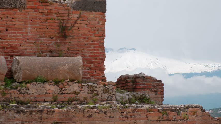 A Slow Rightward Pan Frames Snowy Mount Etna Between the Weathered Brick Walls and Stone Fragments of Teatro Antico di Taormina in Sicily.