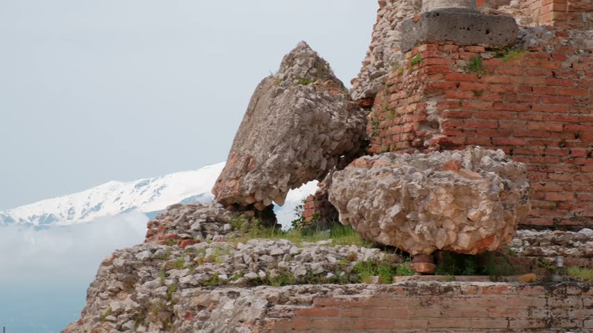 A Leftward Pan Crosses Stone Fragments and Brick Walls at Teatro Antico di Taormina, Briefly Framing Snow-Covered Mount Etna Between the Ruins.