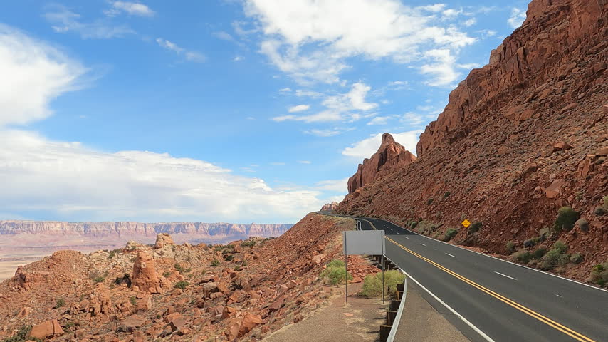 A scenic paved road winding through a red rock canyon landscape under a blue sky with clouds