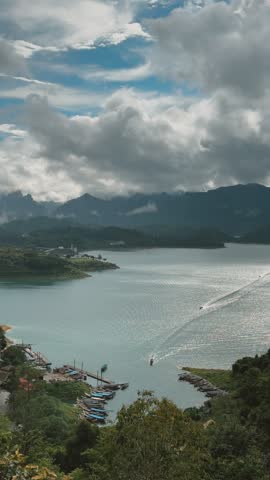 Elevated view of Cheow Lan Lake, surrounded by lush green mountains under a cloudy sky. Boats are docked along the shore.