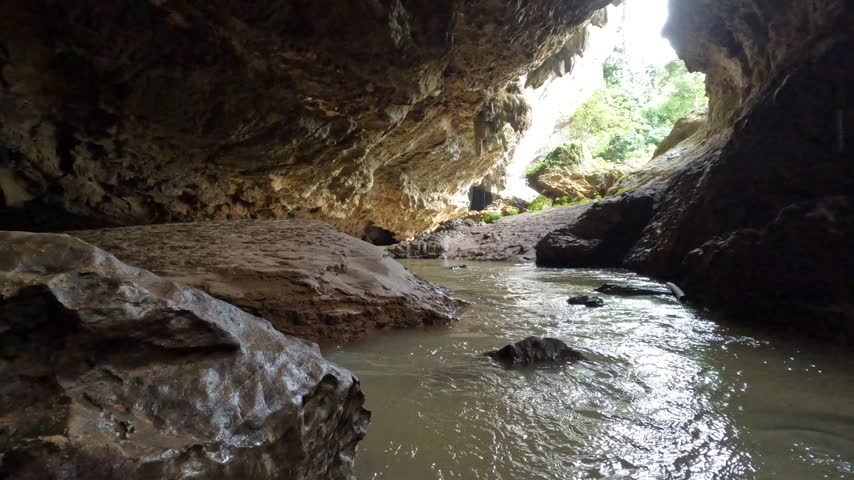 A natural stream flows through a large, dark limestone cave, illuminated by light from the distant entrance. Rugged rock formations define the serene underground.
