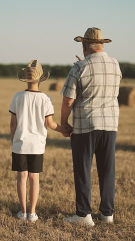 Old farmer and his little grandson admiring beautiful rural landscape in summer, back view. Vertical shot with picturesque scenery, child spending summer vacation in farm of his grandfather, family