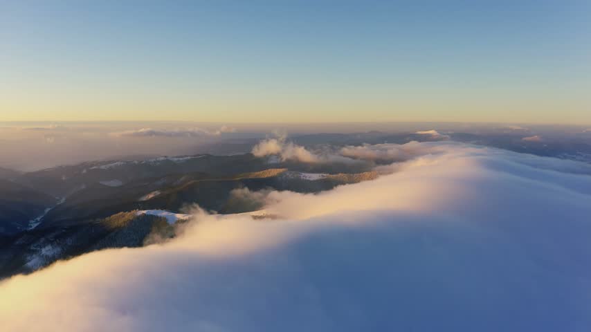 Cloud waves gently and slowly flow over the tops of snow-capped mountains covered with spruce forests at a cold sunset