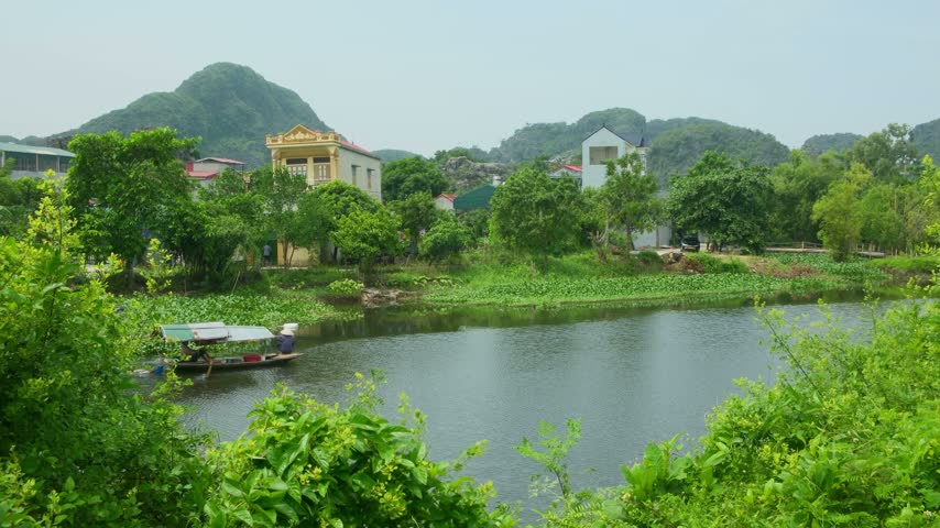 Traditional wooden fishing boat crossing a river through a small rural village in Ninh Binh, Vietnam, surrounded by countryside, vegetation, and local homes.