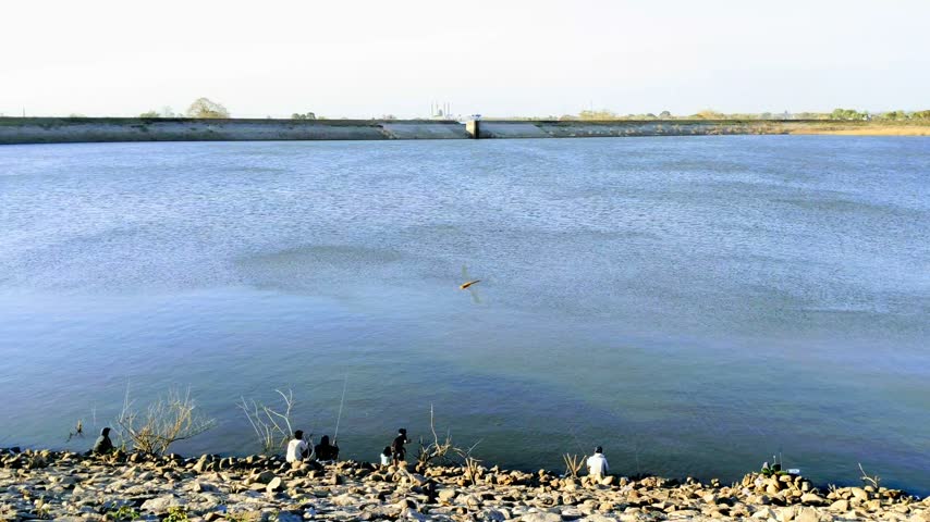 Wide static shot of a vast reservoir lake with people fishing on the rocky embankment under a clear blue sky.