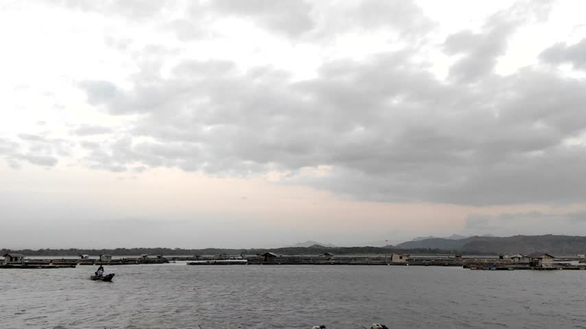 Wide landscape view of a calm reservoir lake with floating fish cages and a moving boat under an overcast evening sky.
