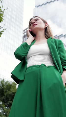Successful female executive wearing a stylish green suit having a serious phone conversation while walking in a modern city district with skyscrapers and trees in the background, low angle view