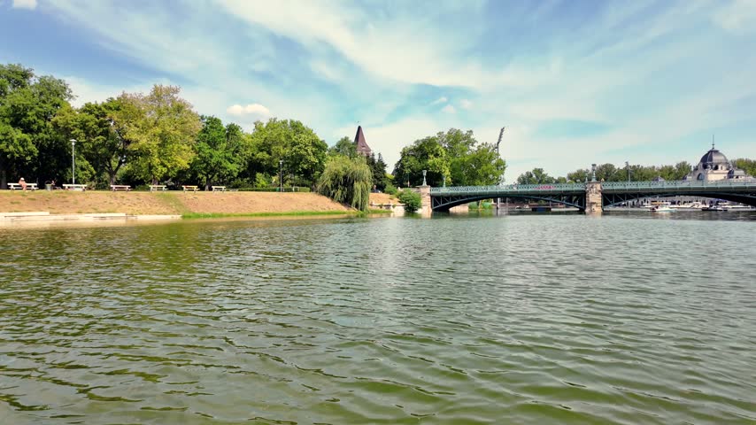 A picturesque view of the Vajdahunyad Castle bridge spanning a lake under a blue sky with clouds. Boats are visible on the water.