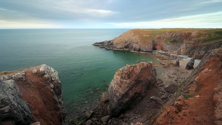 Rocky foreground and steep coastal cliffs frame a peaceful sea view along the rugged Welsh coastline.