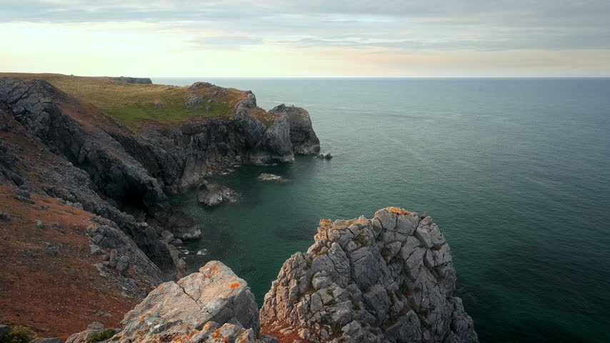 A high coastal viewpoint overlooks steep cliffs, offshore rocks and calm blue water along the Welsh coast.