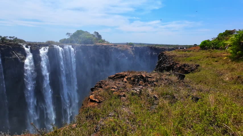 A panoramic view sweeps across Victoria Falls, showing several cascades dropping over dark cliffs beside a dry, grassy rim under a blue sky.