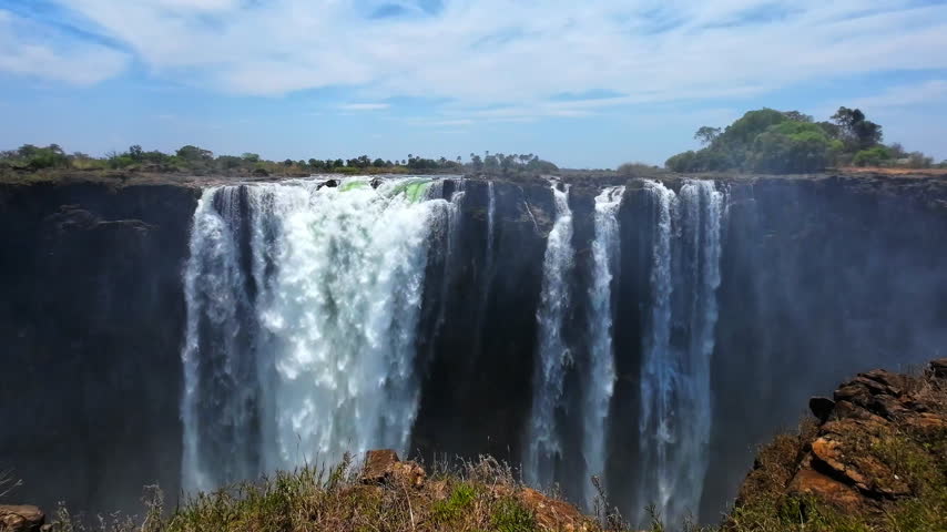 Broad and narrow curtains of water spill over the dark cliff at Victoria Falls, with mist rising from the gorge and trees beyond the riverbank.