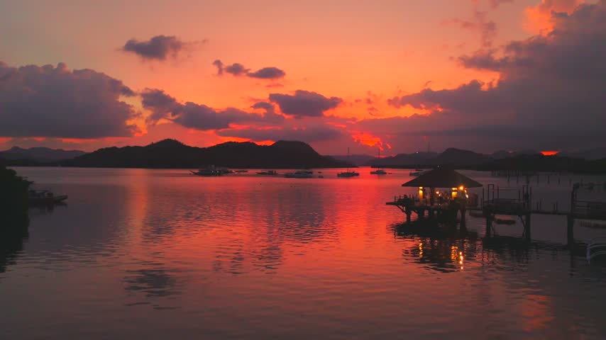 Warm red sunset reflects across Coron Bay beside a lit waterfront pavilion and distant boats