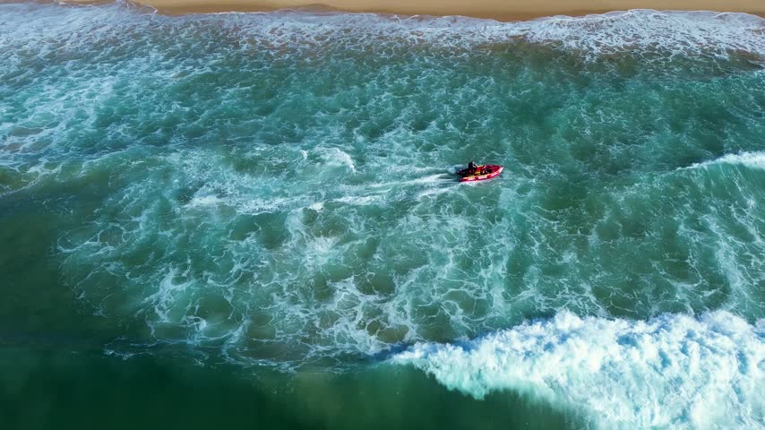 Drone aerial landscape of an inflatable search and rescue lifesaving club patrol boat cruising the shallow beach sandbar with ocean waves crashing on the sandy shoreline on the Central Coast Australia