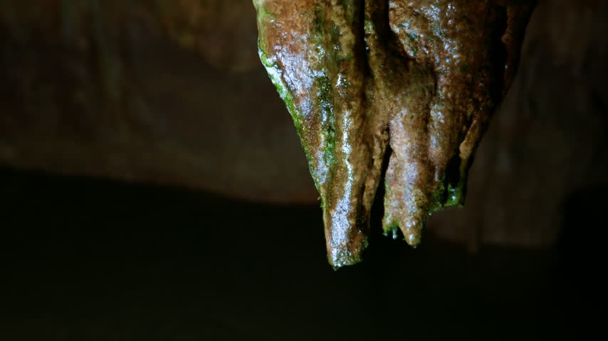 Water droplets cling to and drip from a wet stalactite with brown and green mineral deposits in a dark underground cave. The shot is a static close-up focused on the geological formation.