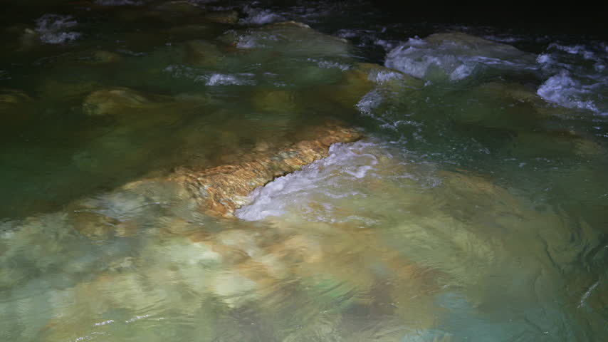 Clear emerald stream flows over textured rock formations inside a dimly lit cavern. Smooth handheld tracking shot follows the current revealing limestone walls and shallow pools.