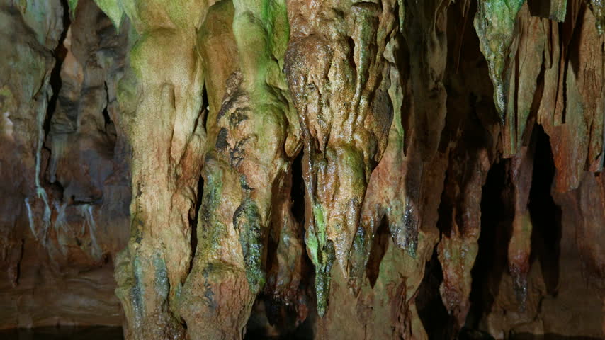 Close-up view of colorful stalactites and stalagmites inside Aggitis Cave, showing green, brown, and beige mineral deposits on rock formations. Static locked shot capturing the textured limestone.