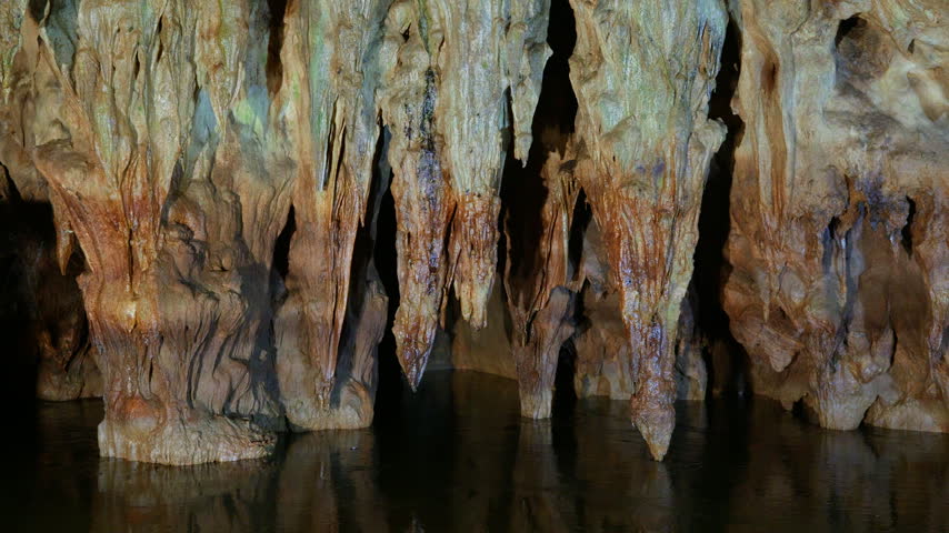 Multiple limestone stalactites with orange and white mineral deposits hang from cave ceiling above dark underground water. Slow lateral pan right reveals intricate rock formations and textured.