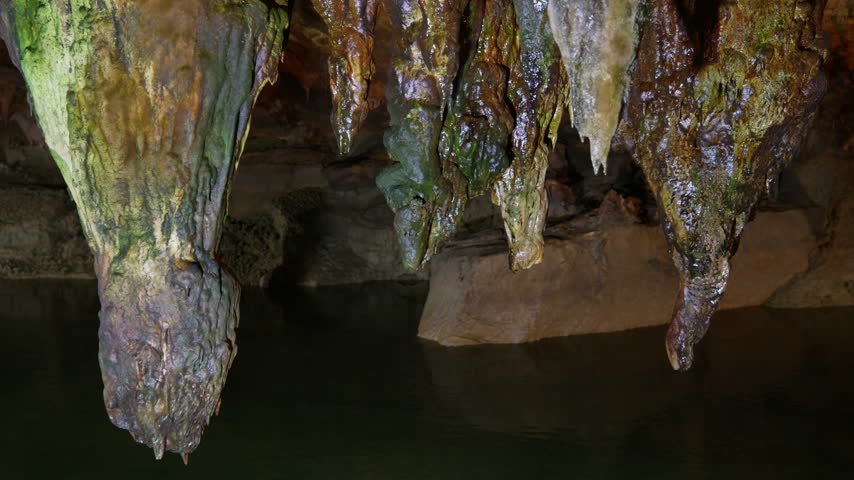 Multiple stalactites hang from a cave ceiling, displaying mineral deposits in brown, orange, and green tones with moss growth. The camera maintains a static upward angle capturing.