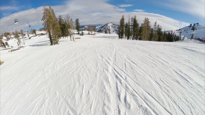 First-person POV of snowboarding down a groomed ski run at Palisades Tahoe on a sunny blue bird day, with scenic mountain terrain surrounding the resort