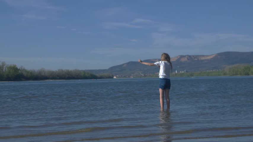 A medium static camera shot captures a young girl in shallow water picking up a rock from the riverbed and throwing it into the Danube river against scenic mountain hills on a sunny day in Hungary.