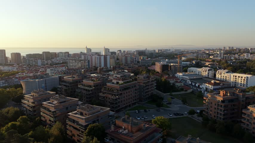 Aerial view of city at sunrise. Buildings glow warmly under soft morning light. Trees line streets, adding green contrast. Sky clears to bright blue, distant sea glimmers. Urban landscape calm