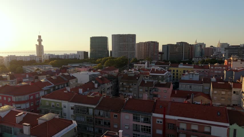 Sunset over Lisbon, Portugal. Red-tiled rooftops stretch toward modern high-rises. Tower rises above urban skyline, bathed in golden light. Warm tones create peaceful, inviting cityscape