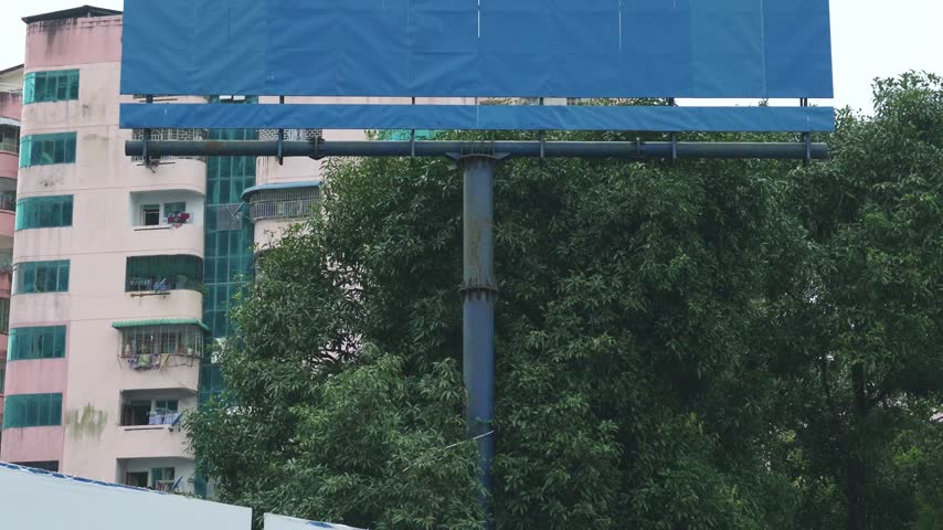 Tilt up shot reveals a large, blank blue out of home advertising billboard standing prominently against a cloudy sky in an urban Yangon neighborhood