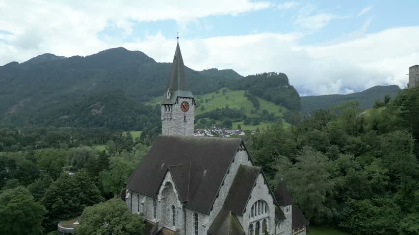 Aerial view of a stone church with a tall steeple nestled among lush green hills and mountains in Liechtenstein.