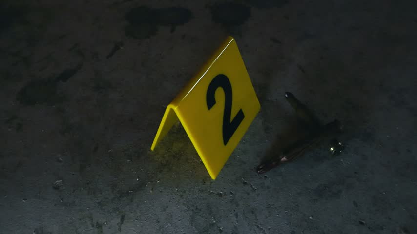 Close up of a yellow evidence marker with the number two next to high caliber bullets on a dark, dusty concrete floor, representing a crime scene investigation or forensic analysis