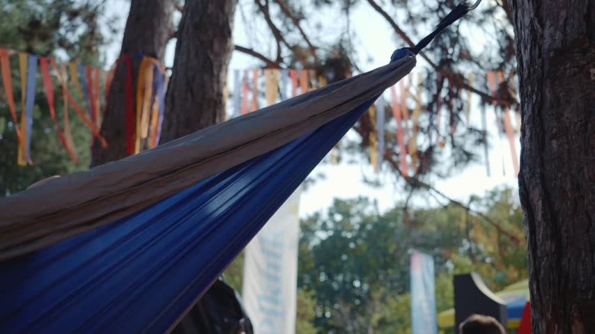 A fabric hammock sways gently between pine trees in an outdoor setting. Colorful ribbons and wooded surroundings are visible in the background.