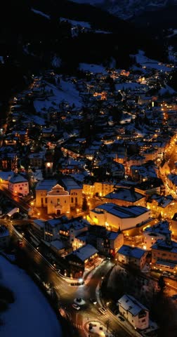 Aerial view of picturesque mountain town illuminated at night with snow covered rooftops and winding roads in winter.