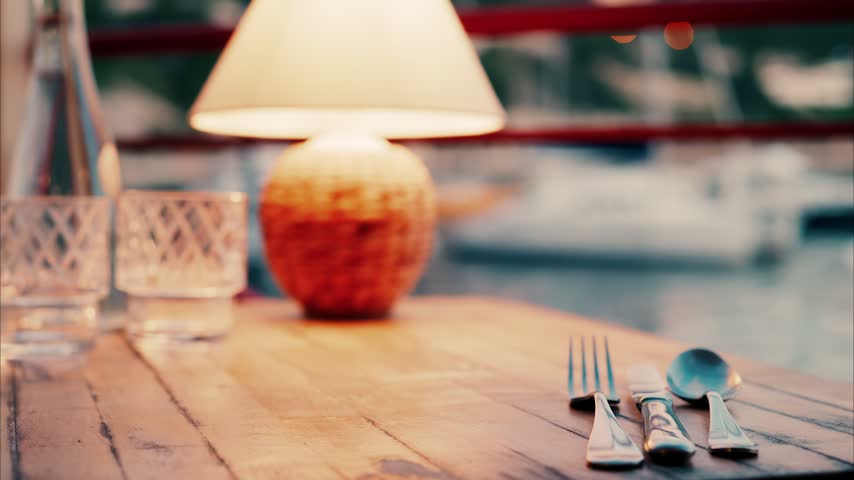 Romantic table setup featuring silverware glassware and soft lamp lighting on wooden surface with blurred boats in harbor background.
