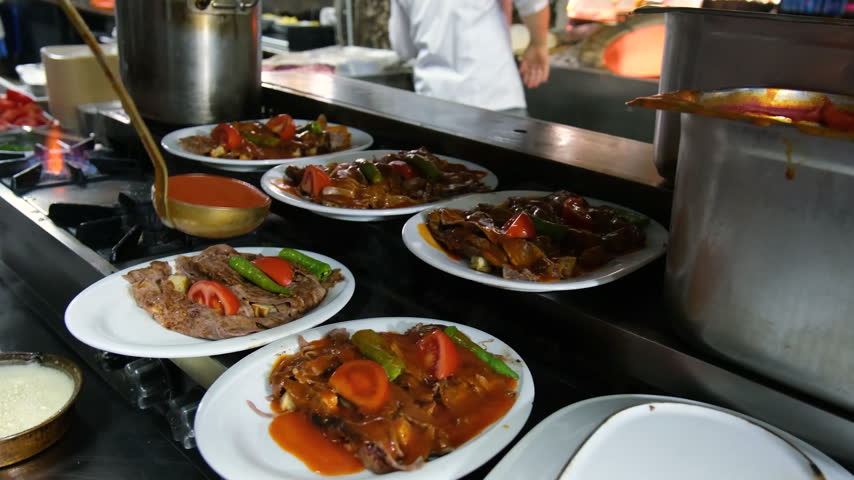 Chef pouring rich tomato sauce over plates of traditional doner kebab in a professional restaurant kitchen with warm natural lighting and close-up perspective.