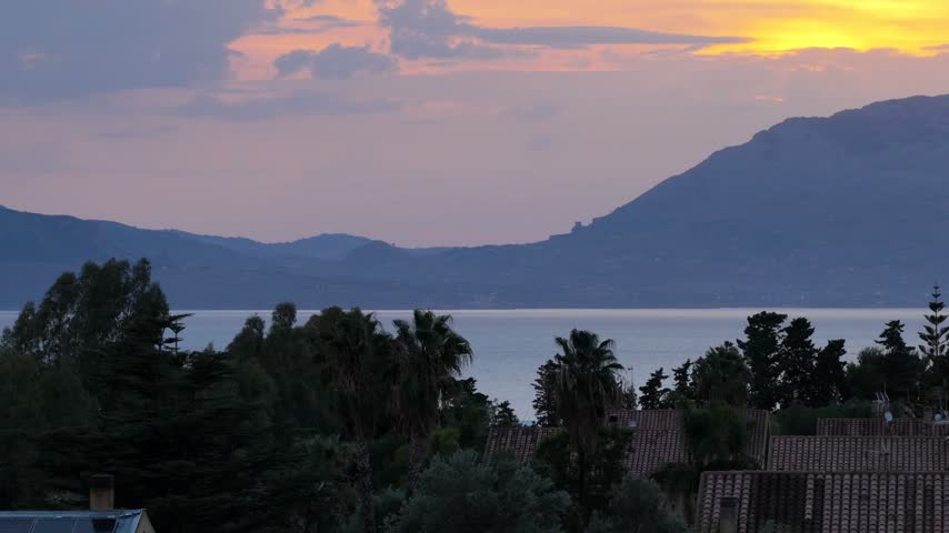 Aerial view of a vibrant sunset over the sea with distant mountains, palm trees, and rooftops in the foreground in Terrasini, Sicily, Italy.