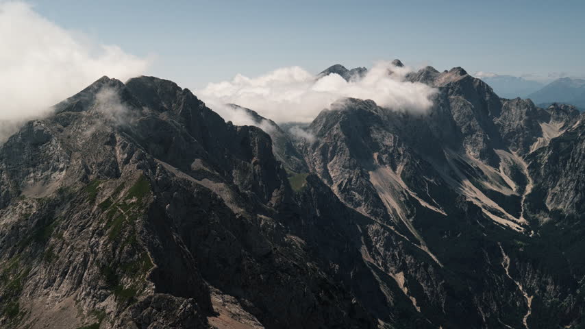 Clouds drift between steep rocky ridges in the Kamnik Savinja Alps, Slovenia. An elevated view looks across deep valleys and rugged mountain slopes.