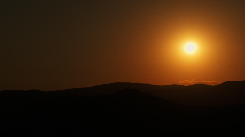 A bright sun hangs above dark hills in a deep orange sky. The steady view shows a thin band of clouds near the horizon and warm evening light.