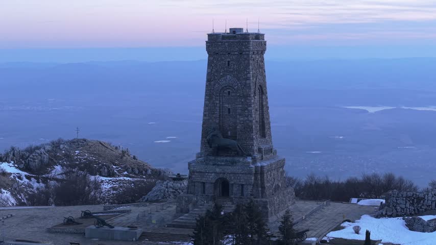 Aerial view of the historic stone Shipka Monument standing on a mountain peak at dusk with snow patches and surrounding valley landscape in Stara Zagora, Bulgaria.