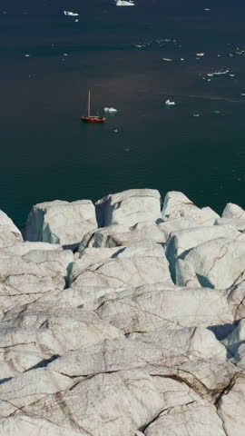 Sailboat Navigating The Dark, Icy Waters Near A Massive Glacier Edge In Scoresby Sound, Greenland. - aerial vertical shot
