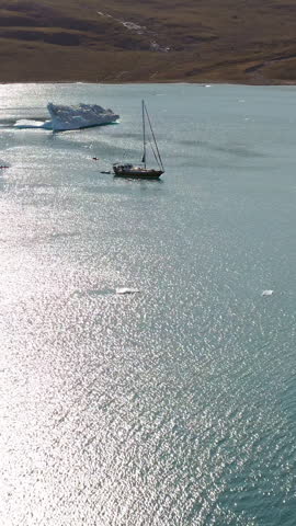 Sailboat Anchored In Calm And Glistening Polar Fjord Waters Near A Floating Iceberg In Scoresby Sund Greenland. - aerial vertical shot
