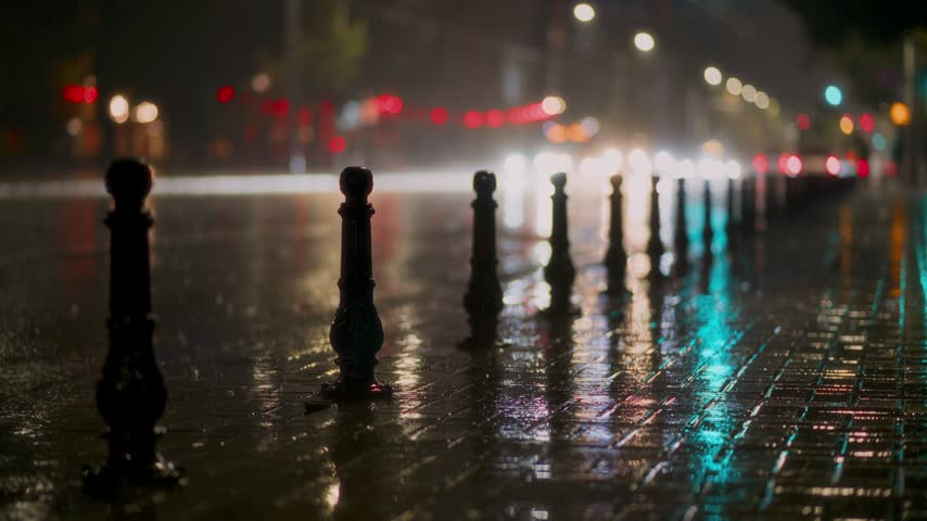 Heavy rainfall on urban road at night featuring blurred car headlights and bokeh city lights on wet cobblestone sidewalk surface.