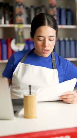 Young latina female employee working at the counter of an auto parts store, checking a list and thinking