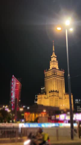 Night view of Palace of Culture and Science in Warsaw with street traffic. Illuminated historic building stands next to modern skyscrapers. Cars drive past on a road near this famous landmark. Red neon glows on a curved residential tower. Vertical video.