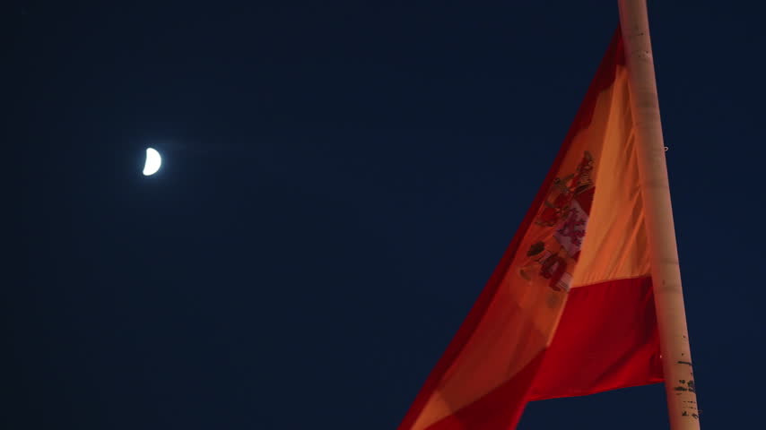 Low angle perspective shows official Spanish national flag waving on flagpole against dark night sky with bright crescent moon in Madrid capital, European political and state news b-roll footage.