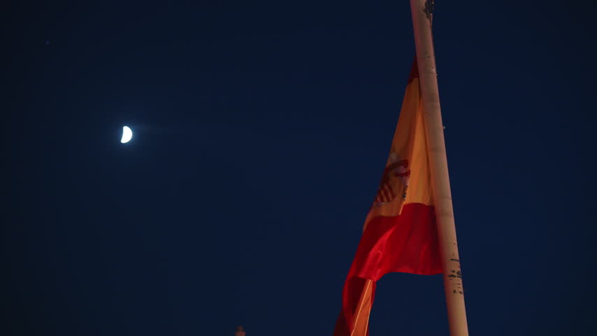 Telephoto close-up shot of Spanish national flag fluttering in wind with illuminated moon in dark night sky over Madrid capital, European culture, history and state identity b-roll footage.
