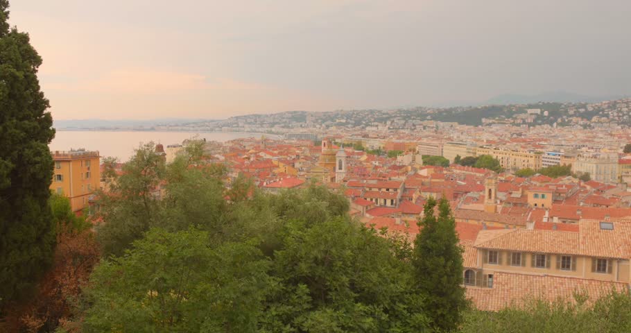 Elevated panoramic view of Nice, France, featuring historic buildings, red tiled rooftops, green trees and the Mediterranean coastline beneath a warm pastel evening sky.